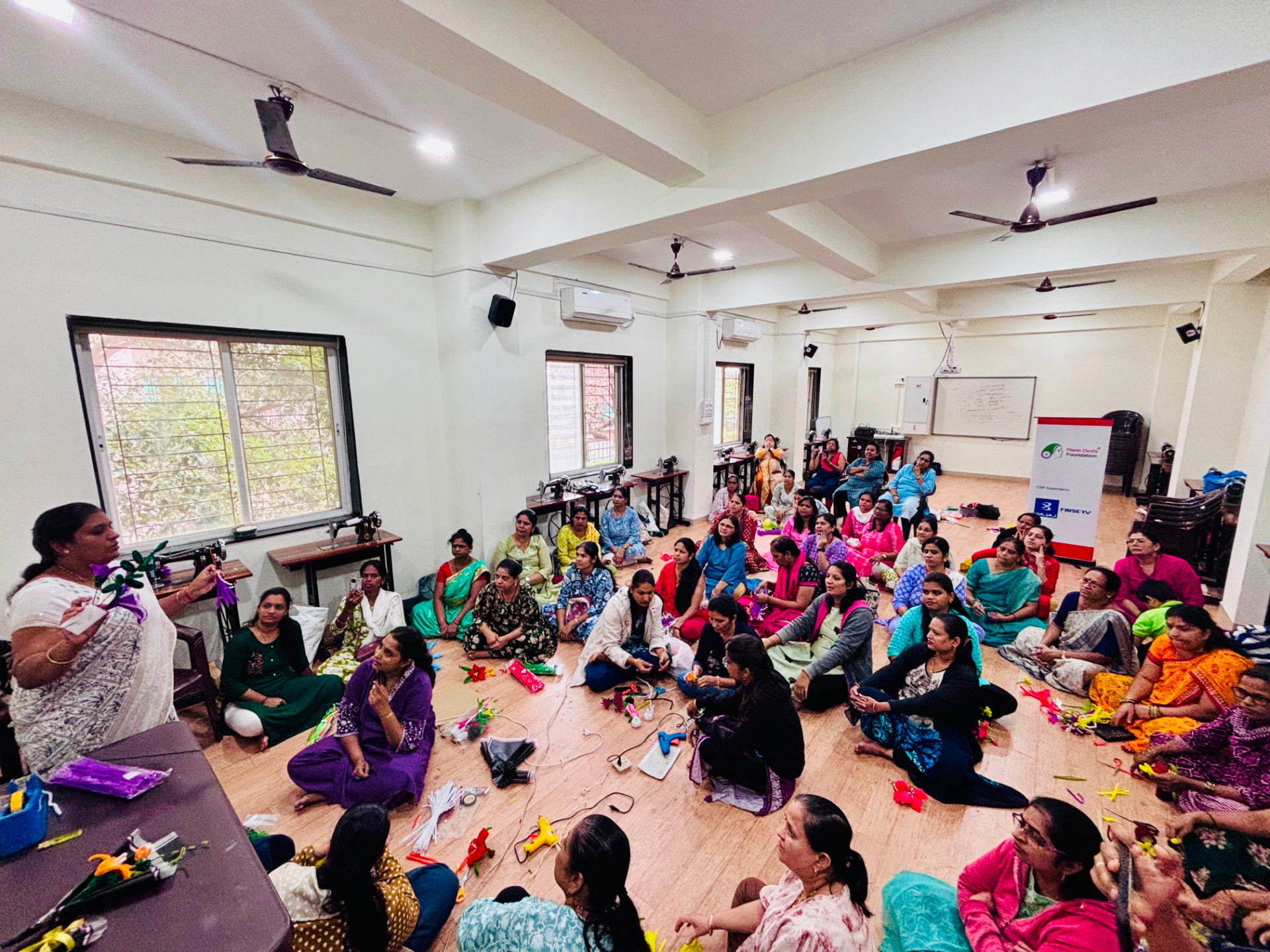 Trainer demonstrating flower making to women seated on the floor of the training hall