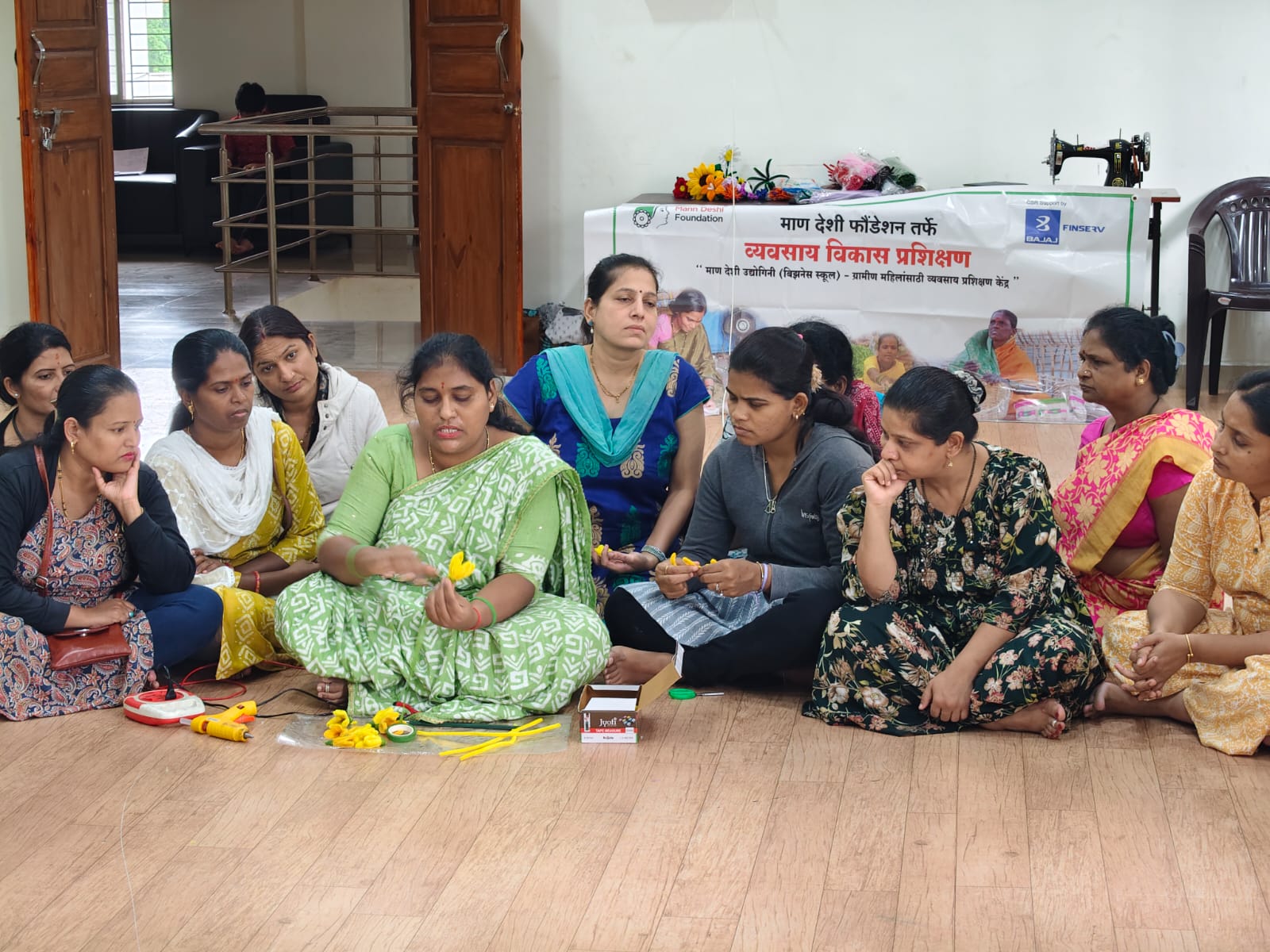 Women seated on the floor making flowers beneath a business development training banner